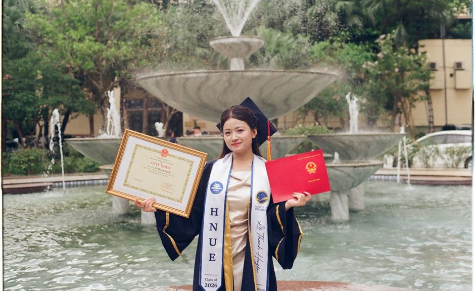A young woman in graduation attire celebrates by a fountain with certificates outdoors.